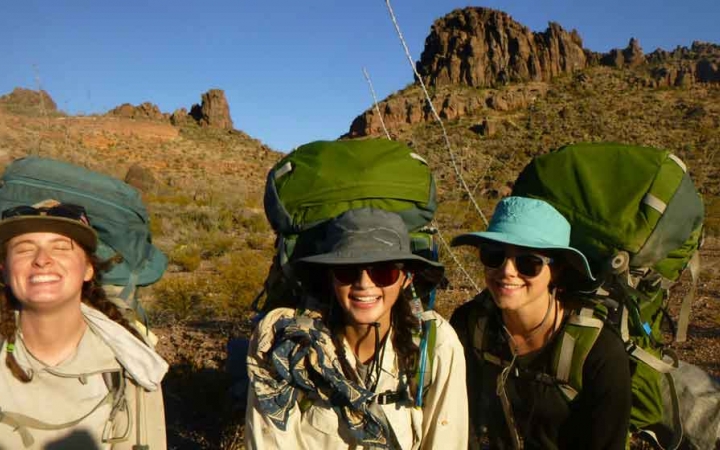 three gap year students wearing backpacks smile at the camera in front of a tall desert rock formation on an outward bound course